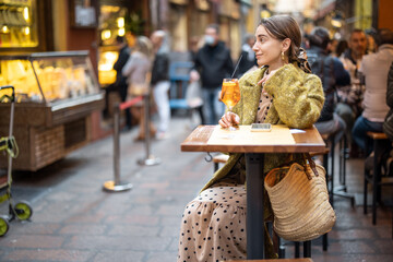 Woman sitting with a cocktail at outdoor bar or restaurant on the crowded street in Bologna....