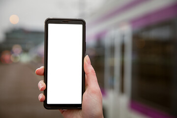 Close-up smart phone with white screen in hand. Railway station platform. Person traveling and using phone app. Trains on the background.