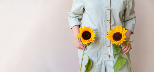 A girl in a light green cotton jumpsuit holds two sunflowers in her hands, a beige monochrome background. The concept of an ecological lifestyle and clothing. copy space