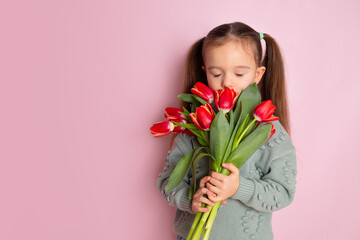 A little cute girl holding a bouquet of tulips and sniffing them with closed eyes Pink background. Happy women's day. Space for text. Bright Emotions.