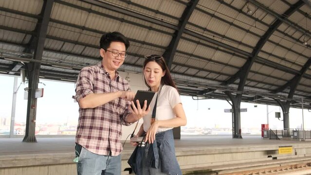 Young couple Asian tourists search information, find travel locations by tablet map at a train station junction in Thailand, passenger holiday trip lifestyle, casual transportation, journey vacation. 