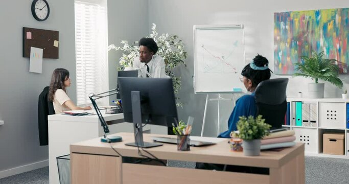 A man with afro hair dressed in a white shirt with a tie leans against a co-worker's desk, asks her to draw a graph on the company's profit projections board