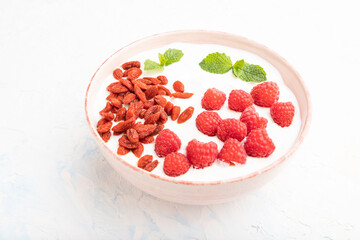 Yogurt with raspberry and goji berries in ceramic bowl on white concrete background. Side view.