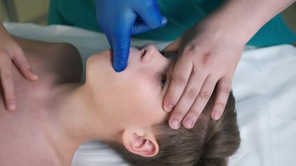 Session of craniosacral therapy, cure of teen boy's jaws by a doctor therapist. Craniosacral therapist in medical gloves makes bite correction by hands at hospital. Boy is lying on couch.
