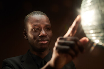 Happy young African man touching discoball and looking at it in the night club while standing in front of camera