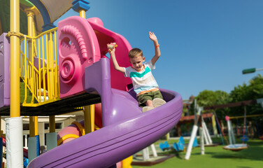 Fototapeta na wymiar On sunny summer day, a boy slides down a slide in a playground in a public park