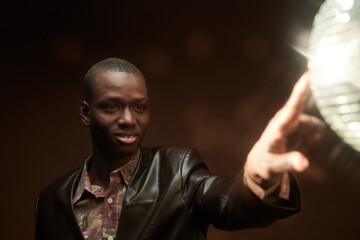 Happy young African man touching discoball at disco in the night club while standing in front of camera over dark background