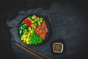 Japanese poke or buddha bowl with salmon, avocado, roe, mango, pies and cucumber in a black bowl on black background, top view with copy space