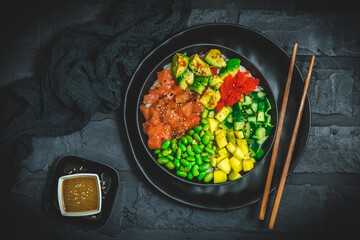Japanese poke or buddha bowl with salmon, avocado, roe, mango, pies and cucumber in a black bowl on black background, top view