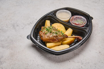 Close up confit duck leg with caramelized apple and celery cream in black plastic container on light background. Designer food concept for take away or delivery
