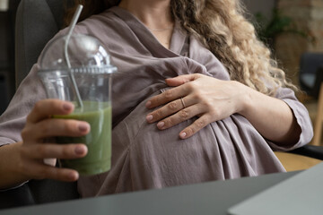 Young pregnant woman in grey dress keeping hand on belly while sitting in armchair by desk and having fruit cocktail or smoothie