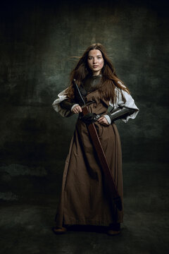 Full-length portrait of seriuos beautiful woman in image of medieval warrior or knight with dirty wounded face with big sword isolated over dark vintage background.