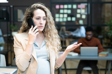 Young confident pregnant businesswoman consulting colleague by mobile phone while standing against coworkers in office