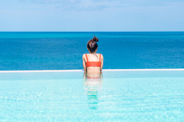 Happy woman in orange swimsuit swimming in infinity pool at luxury hotel against ocean front. young female enjoy in tropical resort. Relaxing, summer,  travel, holiday, vacation and weekend concept