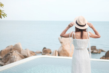 Happy traveler woman in white dress and hat enjoy beautiful sea view, young woman standing at swimming pool. Freedom, relaxing, vacation holiday and summer travel concept