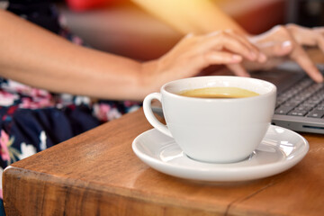 Close-up view, Warm white coffee cup on table desk with blurred woman hand typing on laptop.