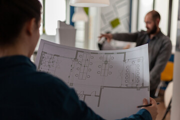 Over shoulder view of architect holding blueprints in architectural office doing teamwork with collegue engineer in the background. Closeup of construction plans of modern business floor plan.