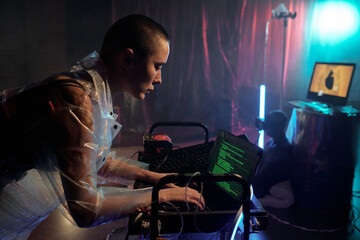 Fototapeta na wymiar Young serious female in cellophane coat decoding data and looking at laptop display against male cyberpunk sitting on the floor