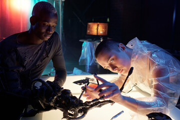 Young female cyberpunk repairing electronic equipment while bending over table illuminated with white light in hacker hideouts