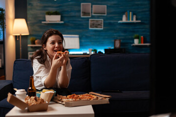 Smiling woman eating a slice of hot pizza delivery sitting on couch looking at family show on...