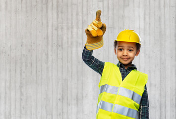 building, construction and profession concept - smiling little boy in protective glove, yellow safety vest and helmet pointing finger up over grey concrete wall background