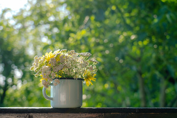 Cute field flowers bouquet. Bunch of wild plants in blue metal mug standing on wooden background isolated on blurry green nature backdrop