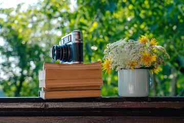 Closeup view stock photography of old vintage photo camera on stack of old paper books on brown wooden table background. Beautiful valley flowers arranged in blue enamel mug