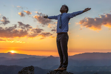 Successful businessman standing raised hand on top of peak mountain golden sunset background,...