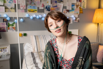 Portrait of teenage girl breathing with special oxygen tube looking at camera while sitting on bed...