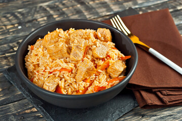 Pilaf with meat and carrots in a black bowl. Pilaf on a black background. Food on a shabby table. Bowl on an old wooden board.