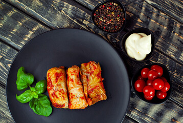 Cabbage rolls with cabbage and rice in a black plate. Food on an old wooden table. Stuffed cabbage on a dark board background. Marinated cherry tomatoes and spices on a plate.