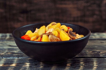 Boiled potatoes with meat in a black bowl. Potatoes with mushrooms and carrots. Boiled mushrooms and chicken with potatoes. Food on a black wooden table.