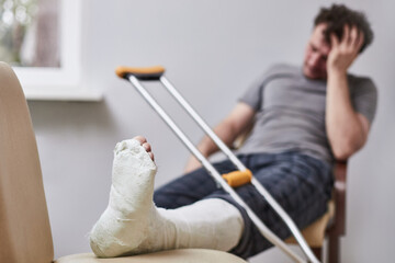 a man with a broken leg and crutches sits grabbing his head. Focus on the foot