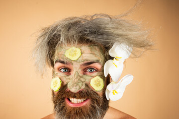 Funny man with clay mask and cucumber slices on face. Man having cosmetic moisturizing mask, isolated studio background. Male face care.