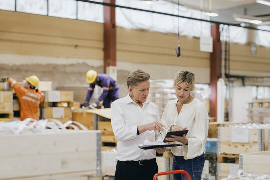 Businesswoman and businessman discussing over digital tablet while workers working in background at warehouse