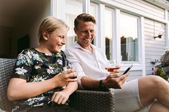 Smiling father sharing smart phone with son while having drinks in back yard