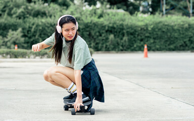 Beautiful sportive hipster happy Asian woman wearing casual shirt and shorts with headphone to listen music while playing skateboard as hobby in free time day on holiday, smiling at outdoor park.