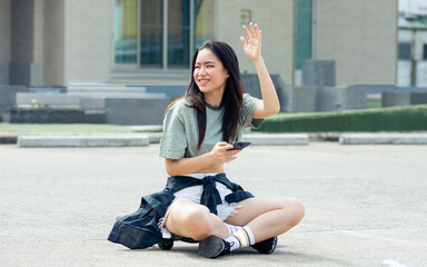 Fototapeta na wymiar Beautiful happy Asian extreme hipster woman smiling, talking mobile phone and waving hand for greeting, sitting on skateboard, relax playing outdoor activity on holidays in summer vacation.