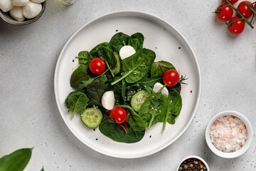 Green organic salad leaves with arugula, spinach, cucumber, cherry tomatoes, mozzarella on white table on grey background.