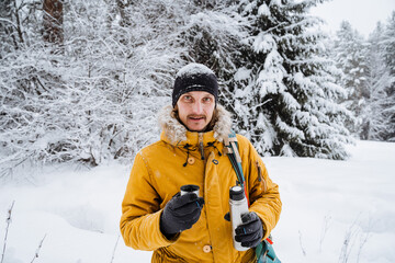 Fototapeta na wymiar Smiling and happy traveler drinks hot tea or coffee from thermos.Snowy forest and rocks. Climbing the mountain in winter. Survival in the wild.