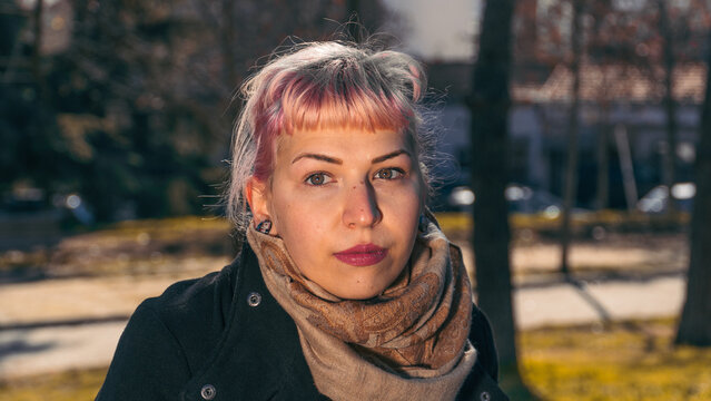 Urbanismo en el estilo propio, chica con flequillo y cabello rosa esperando en un parque, mujer con pelo rosa y bufanda marr&oacute;n en mitad de un parque