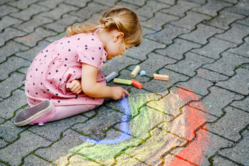 Little preschool girl painting rainbow with colorful chalks on ground on backyard. Positive happy toddler child drawing and creating pictures. Creative outdoors activity in summer.