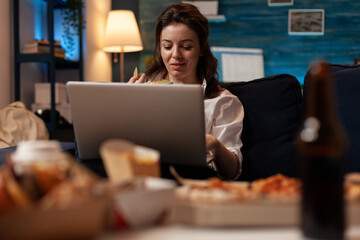 Fototapeta na wymiar Woman browsing social media on laptop while having fast food snack sitting on couch in living room. Office worker with personal computer eating chips in front of table with home delivery menu.