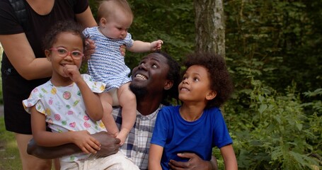 A portrait of a caucasian cute baby girl and smiling African American man with kids in the city park. Big family.