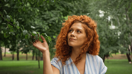 pleased redhead woman touching green leaves on tree.