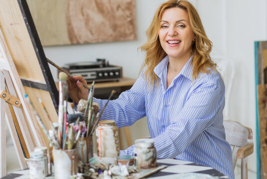 Mature woman painting brush on canvas at studio and looking at camera.