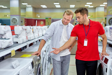 male customer choosing washer machine in shop. difficult decision. Various choice. Pleasant consultant in red uniform help with choice, talking, discussing which option is best. consumerism concept