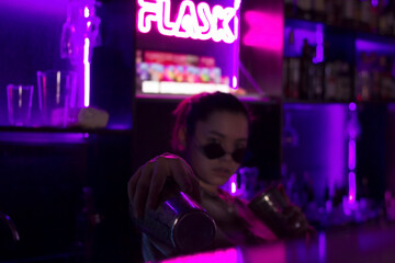Young caucasian woman bartender with cocktail shaker and sunglasses in nightclub bar with red pink and violet purple neon light, barman looking at camera at club party, selective focus