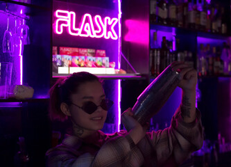 Young smiling caucasian woman bartender with cocktail shaker and sunglasses in nightclub bar with red pink and violet purple neon light, barman at club party, selective focus