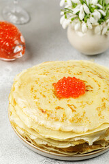 A stack of Russian thin pancakes with red caviar in plate on table. Against background is a vase with snowdrops. Delicious dessert for breakfast.